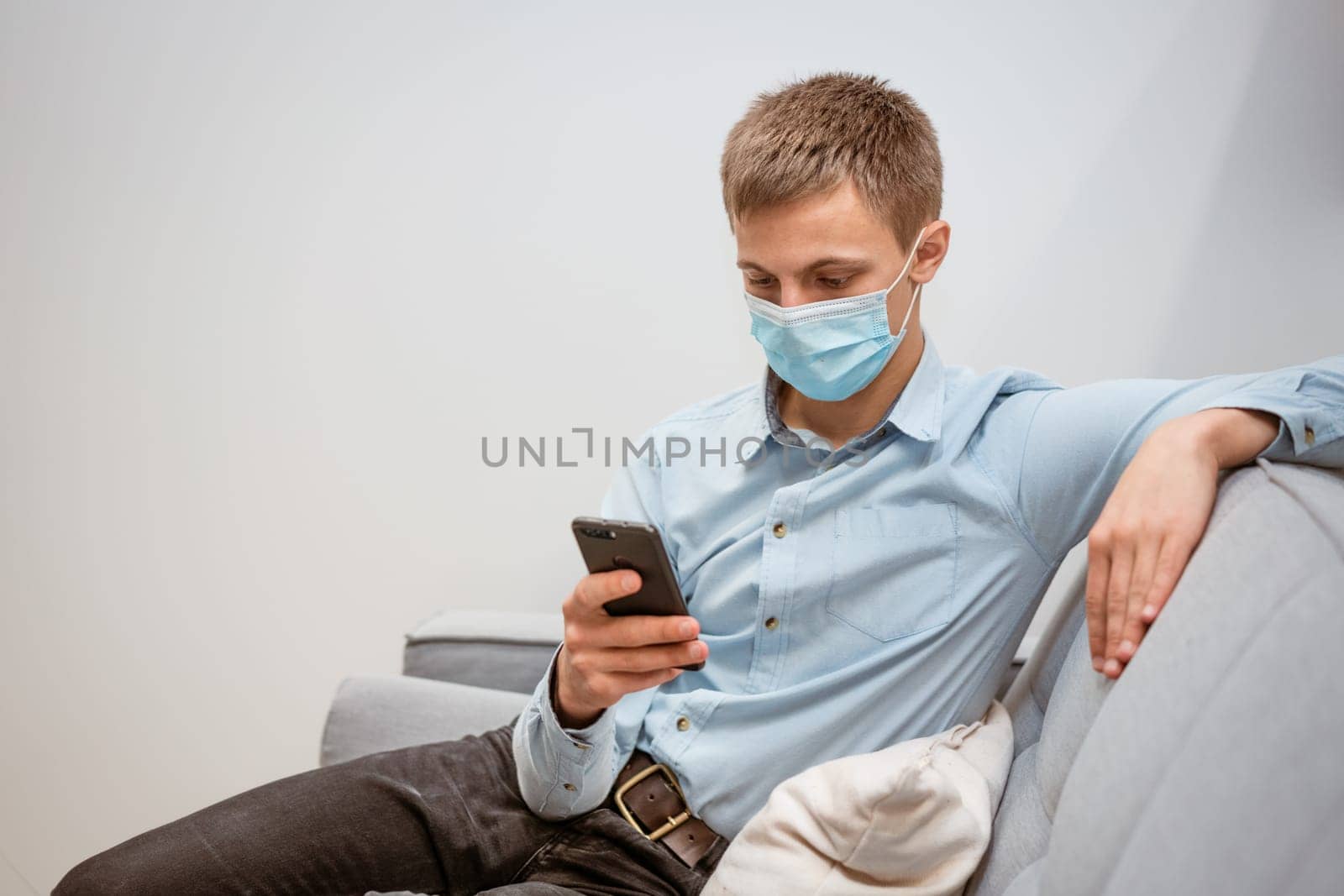 young man of European appearance in casual clothes, at home on the couch in a medical mask communicates by video communication on the phone, the concept of remote online communication on the Internet