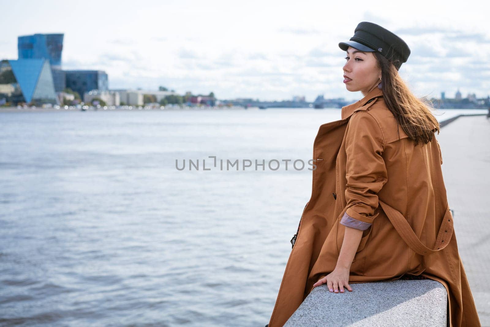 A sad lonely woman of European appearance in a jacket and a cap sits on the embankment of the river on a gloomy day, looks into the distance