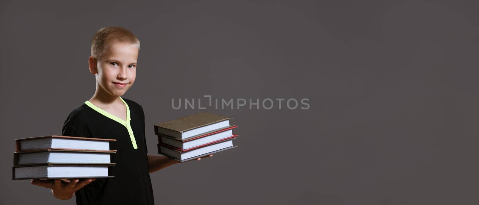 Cute boy in a black t-shirt holds stacks of books on a gray background