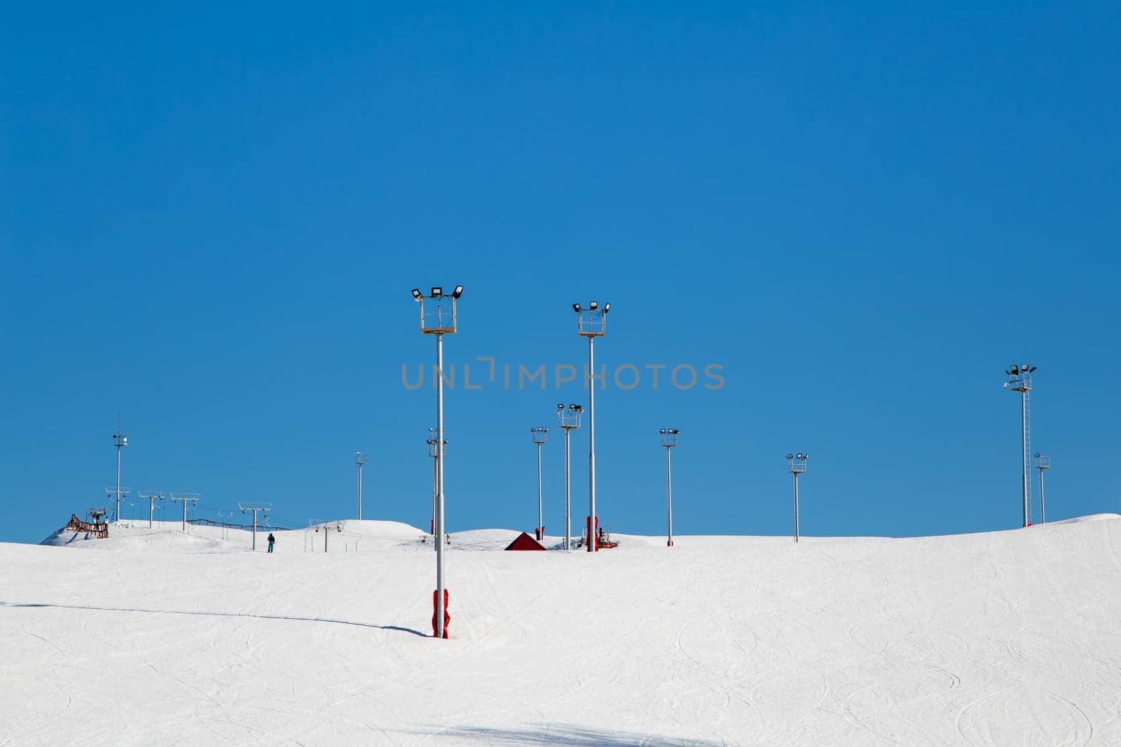 Ski resort, snow slope, trail with artificial lighting towers. Mountain slope for skiing and snowboarding. Against the background of the blue sky. The summit of the rise and ski tours on a sunny day.
