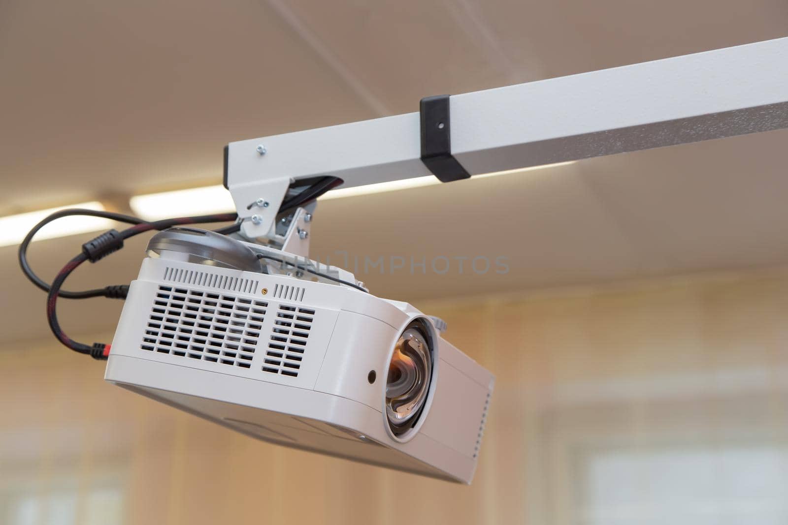The video projector hangs from the ceiling on a white bracket. The fluorescent lamps are on at the back. Window openings and curtains out of focus.