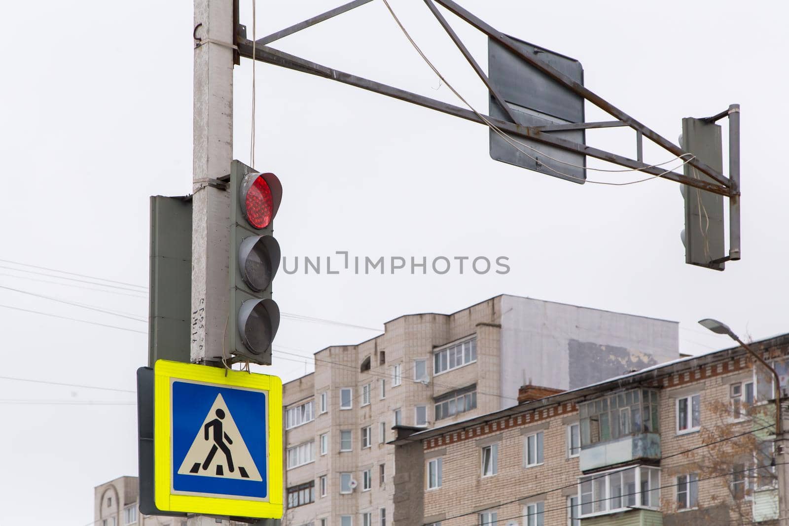 A road sign means a pedestrian crossing, a red traffic light for cars is on. Cars must stop and people can cross the road. Against the background of a gray sky and a residential building.