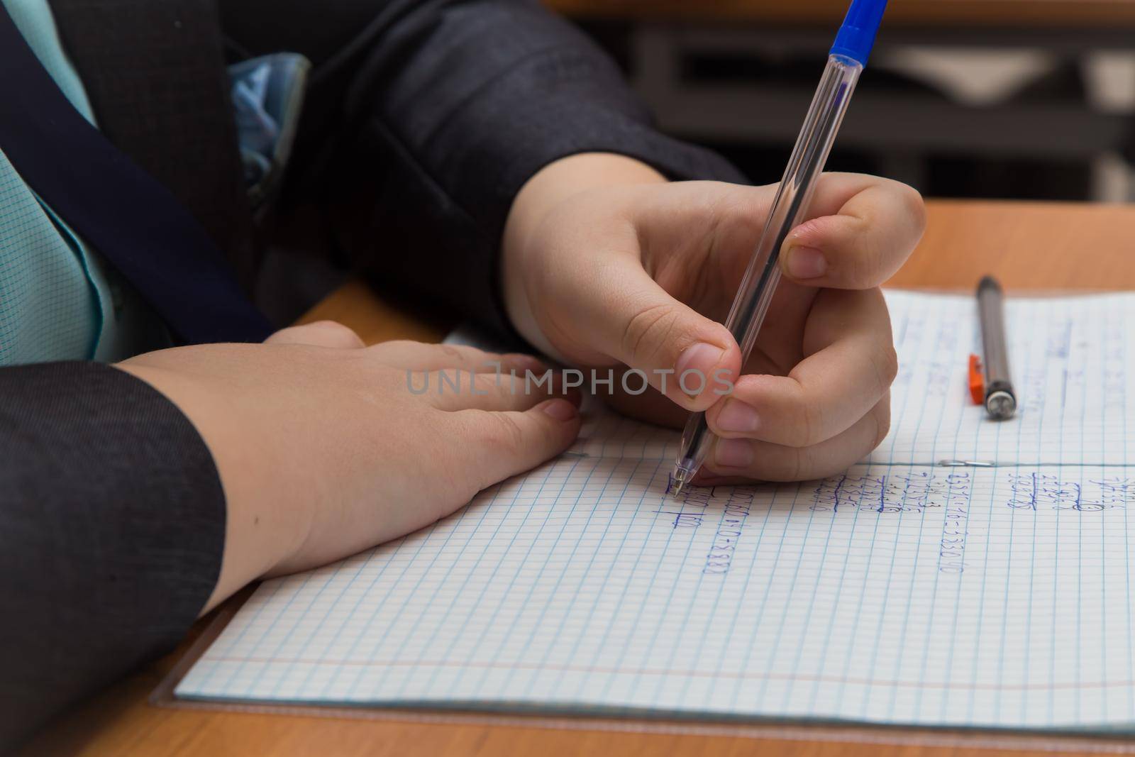 The boys hand with a fountain pen carefully solves a close-up math example. The student completes the task in teradi. A schoolboy teaches lessons at a school desk. Warm soft daylight.