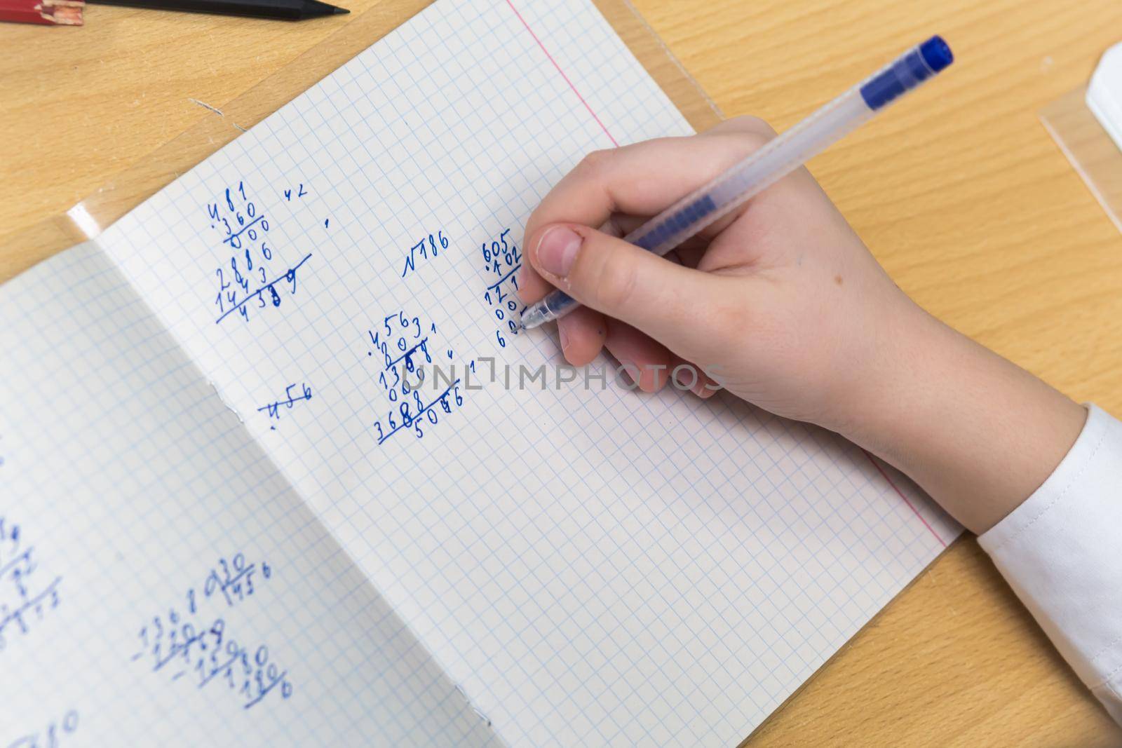 The girls hand with a fountain pen with blue ink solves examples in mathematics. The student completes the task in teradi. A schoolboy teaches lessons at a school desk. Warm soft daylight.