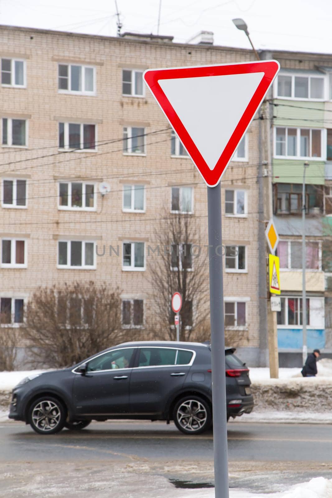 A white triangle sign with a red outline means give way. Against the background of a road with a moving car and a residential building. Winter, white snow lies outside.
