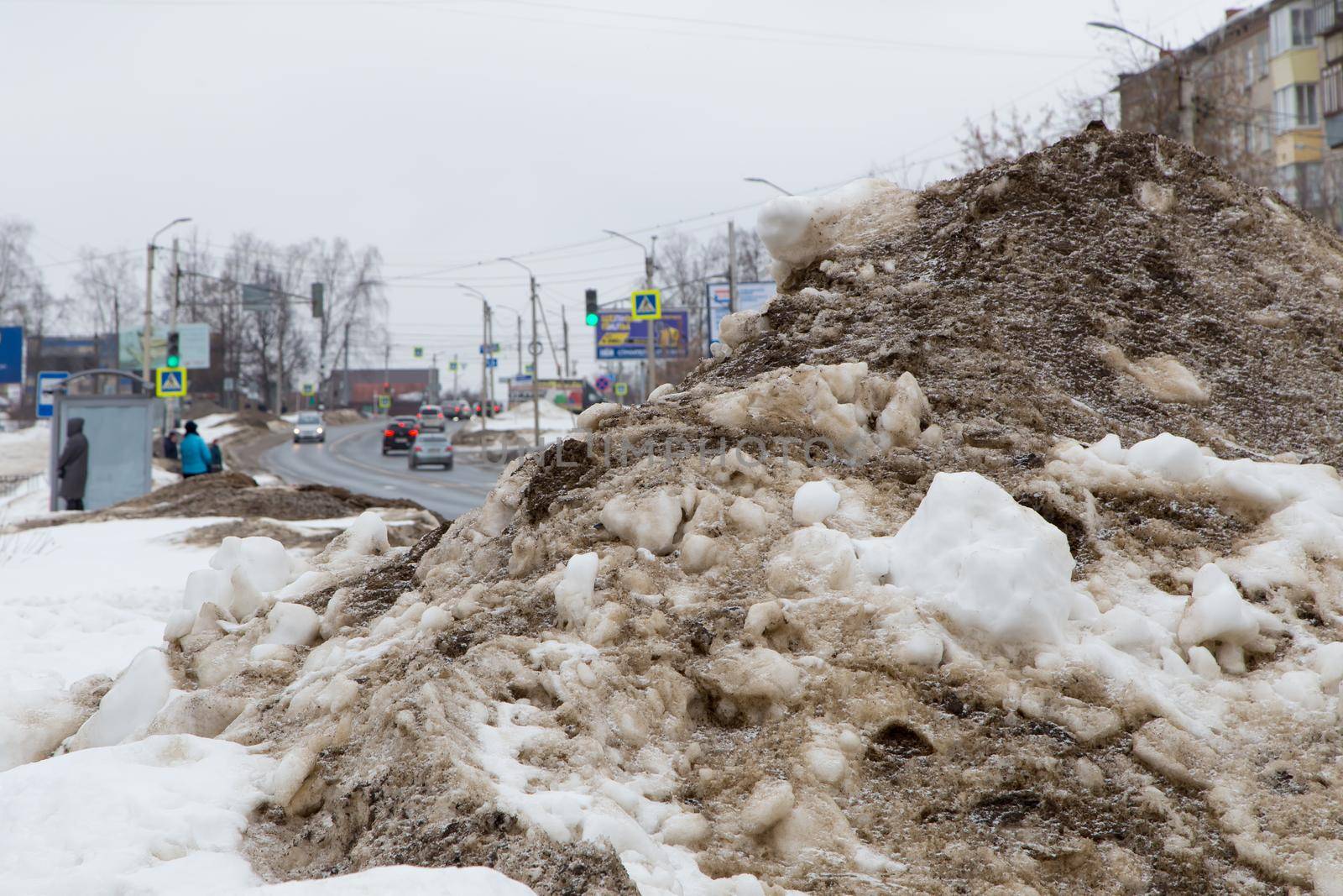 A large pile of snow on the side of the road with passing cars. People at a bus stop, a working traffic light and cars moving along the road. Against the background of a gray cloudy sky.