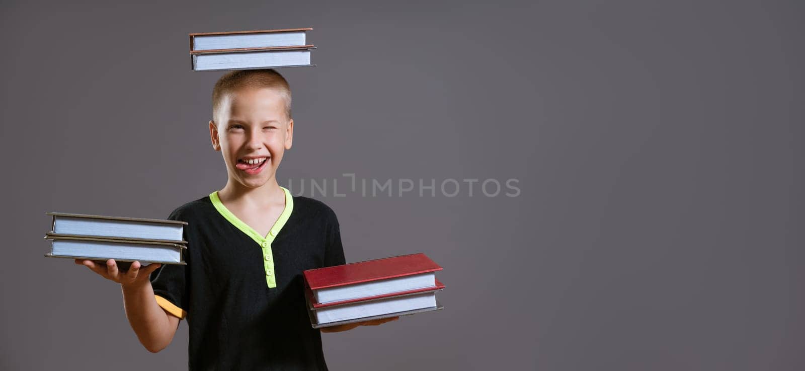 Funny Caucasian boy holding books in his hands and on his head. Shows her tongue on a gray background in casual wear. Back to school concept