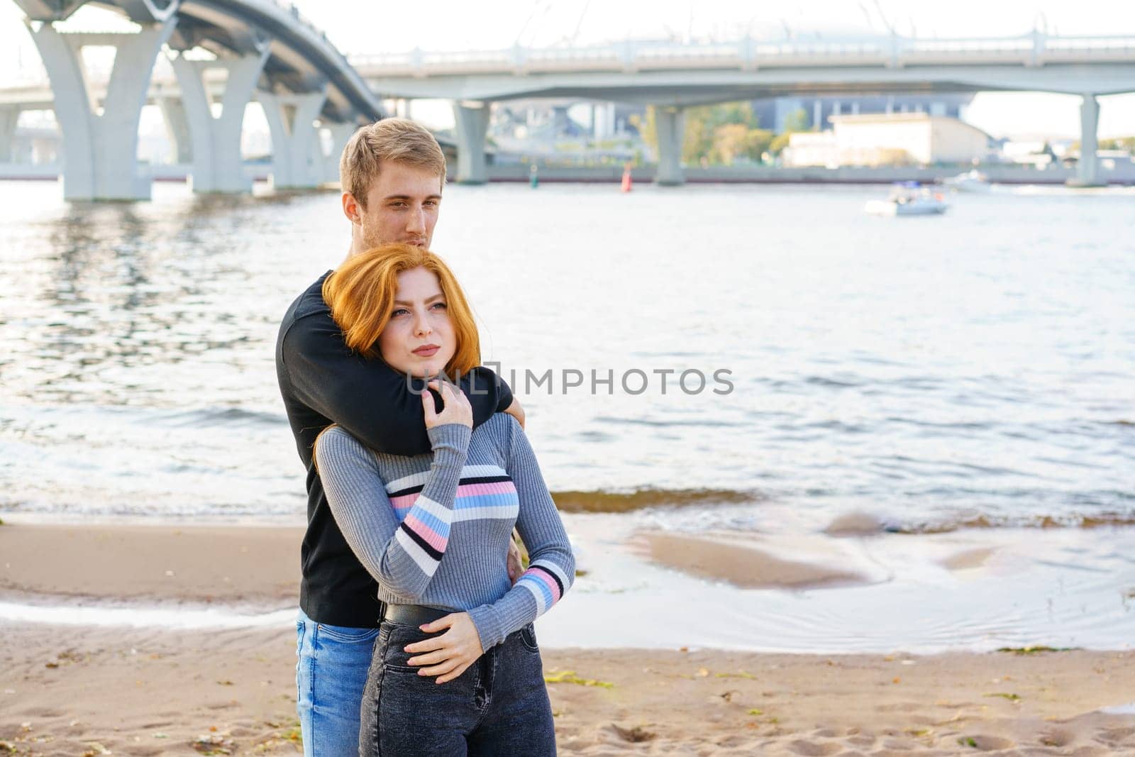 Young couple of man and woman with long red hair of Caucasian ethnicity, in casual clothes, stand on the bank of the river embracing happy on a summer day against the background of the cityscape