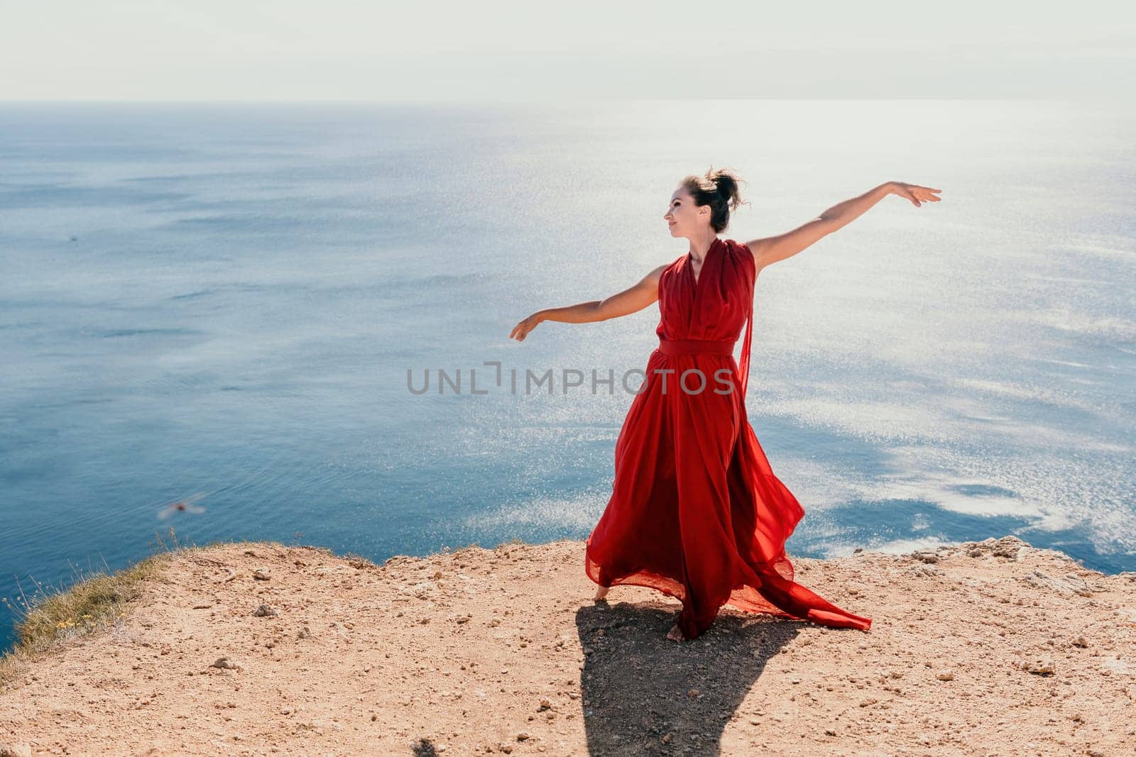 Side view a Young beautiful sensual woman in a red long dress posing on a rock high above the sea during sunrise. Girl on the nature on blue sky background. Fashion photo.