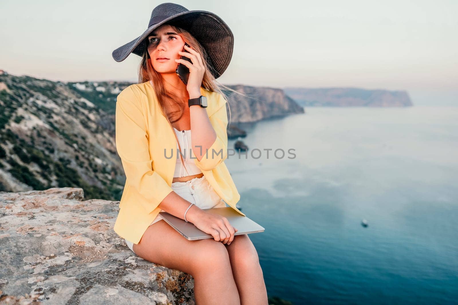 Successful business woman in yellow hat working on laptop by the sea. Pretty lady typing on computer at summer day outdoors. Freelance, travel and holidays concept.