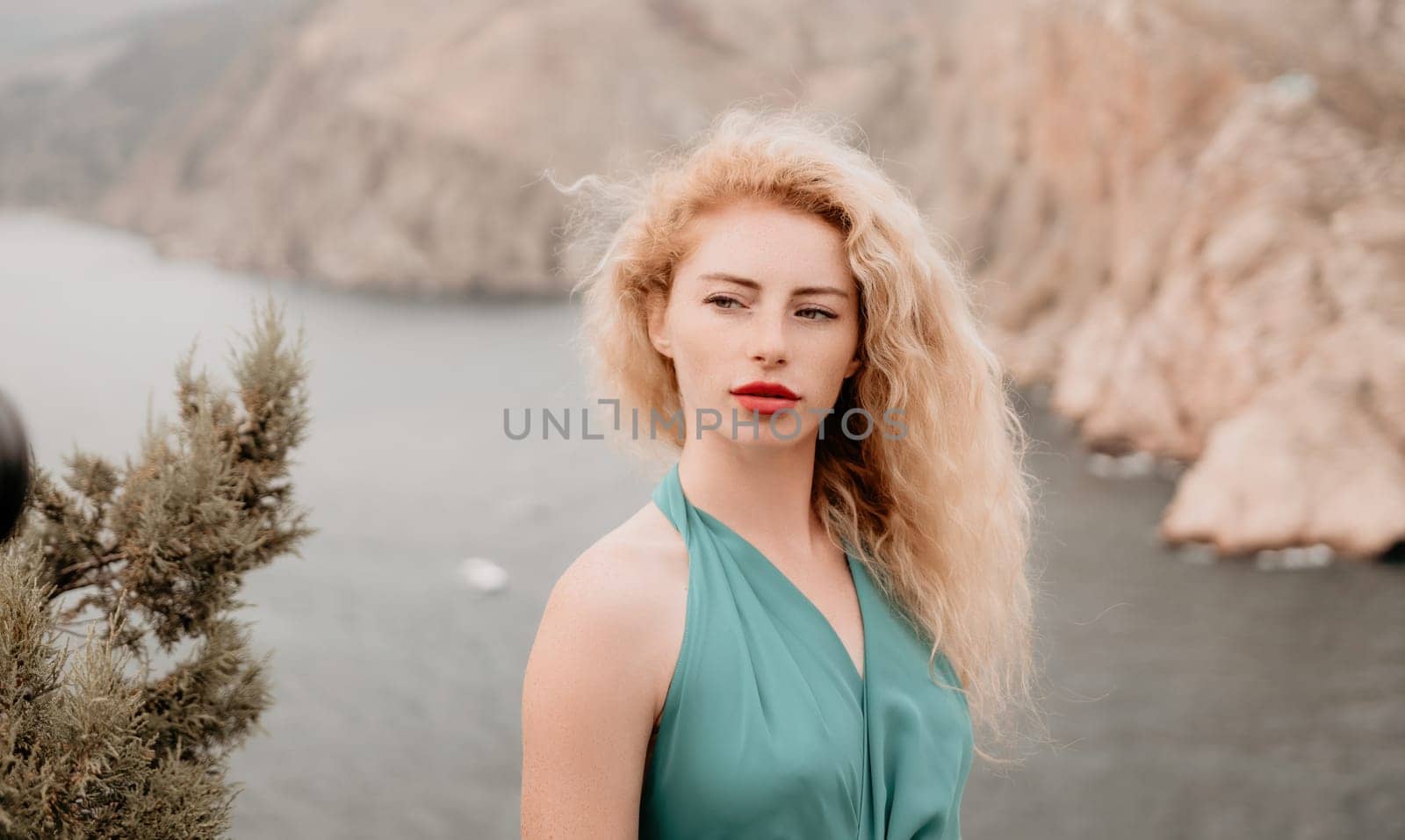 Redhead woman portrait. Curly redhead young caucasian woman with freckles looking at camera and smiling. Close up portrait cute woman in a mint long dress posing on a volcanic rock high above the sea by panophotograph
