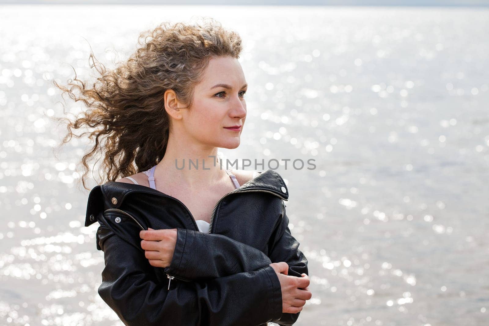 Closeup portrait of a woman, Caucasian woman with curly hair in a black leather jacket looks to the side on the shore against the background of water