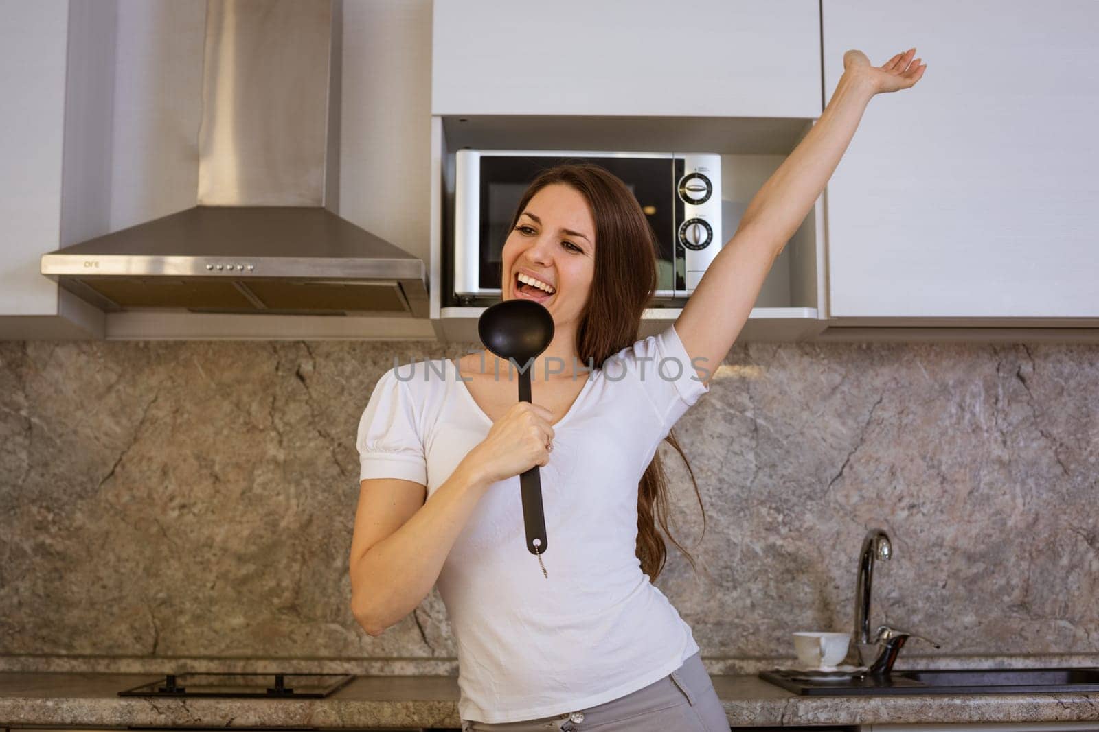 a young brunette woman in a white T-shirt sings in a modern kitchen with a ladle, using it as a microphone