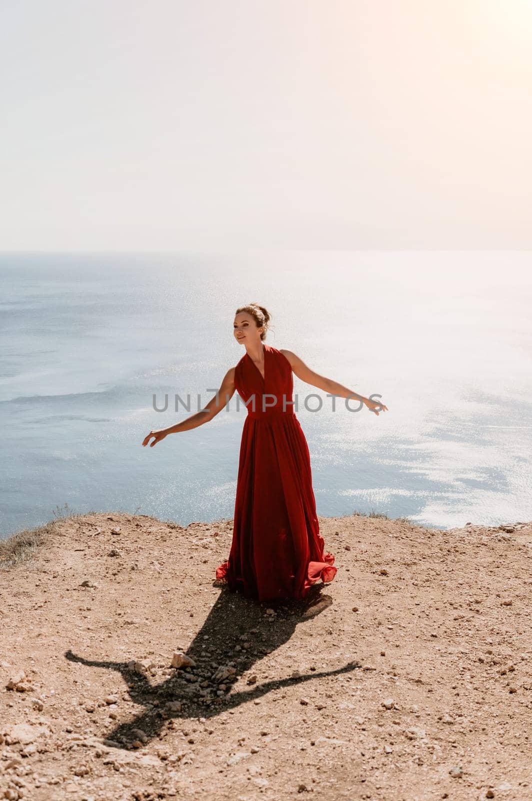 Side view a Young beautiful sensual woman in a red long dress posing on a rock high above the sea during sunrise. Girl on the nature on blue sky background. Fashion photo.