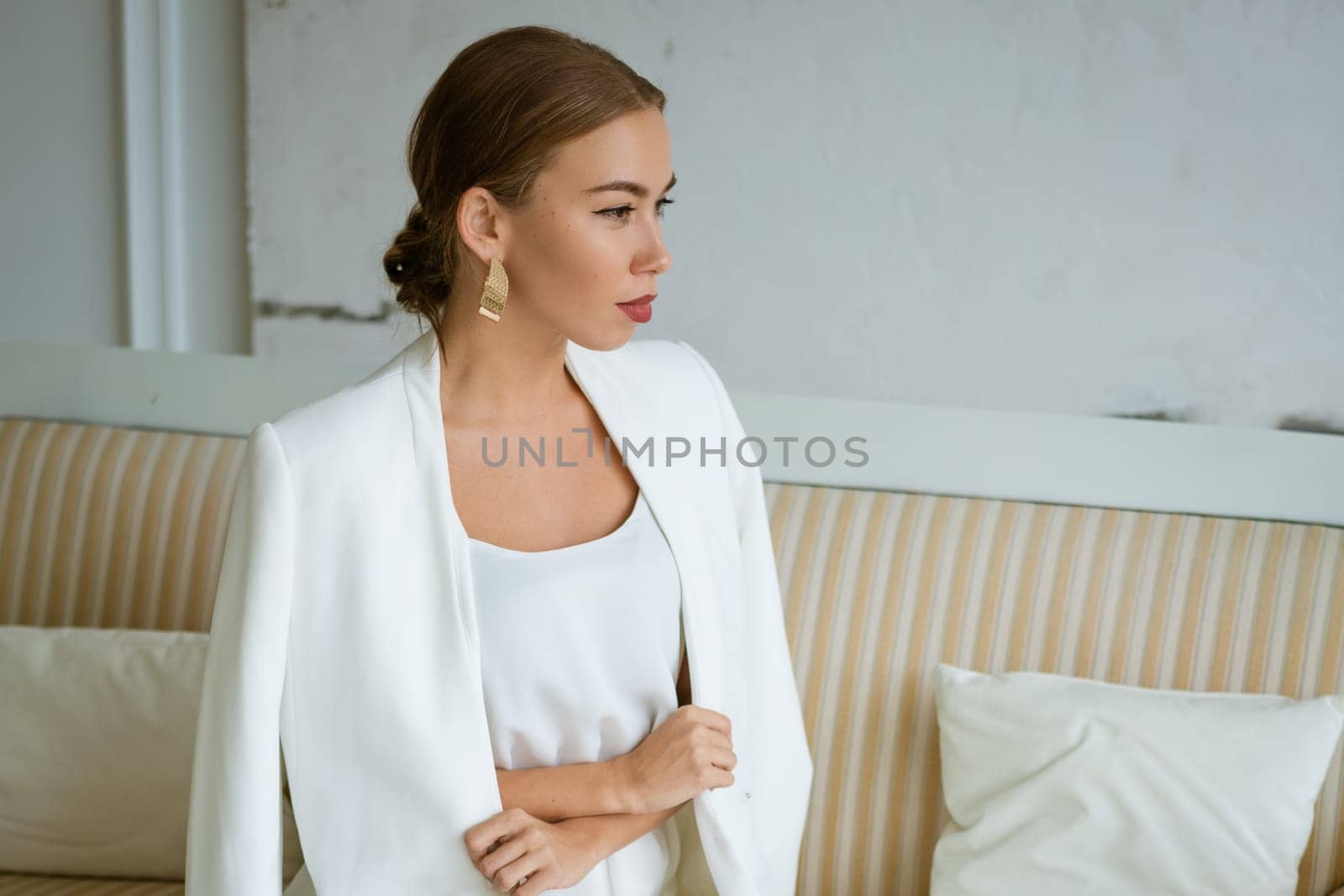 Young business woman of Caucasian ethnicity is sitting on the sofa in white business clothes with beautiful makeup and hair. Portrait of a confident beautiful girl