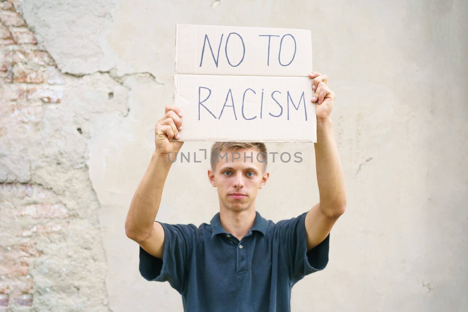 Caucasian guy came out to protest against racism with poster in his hands. Young man appearance stands against background of cement wall with cardboard in his hand on which it is written no to racism