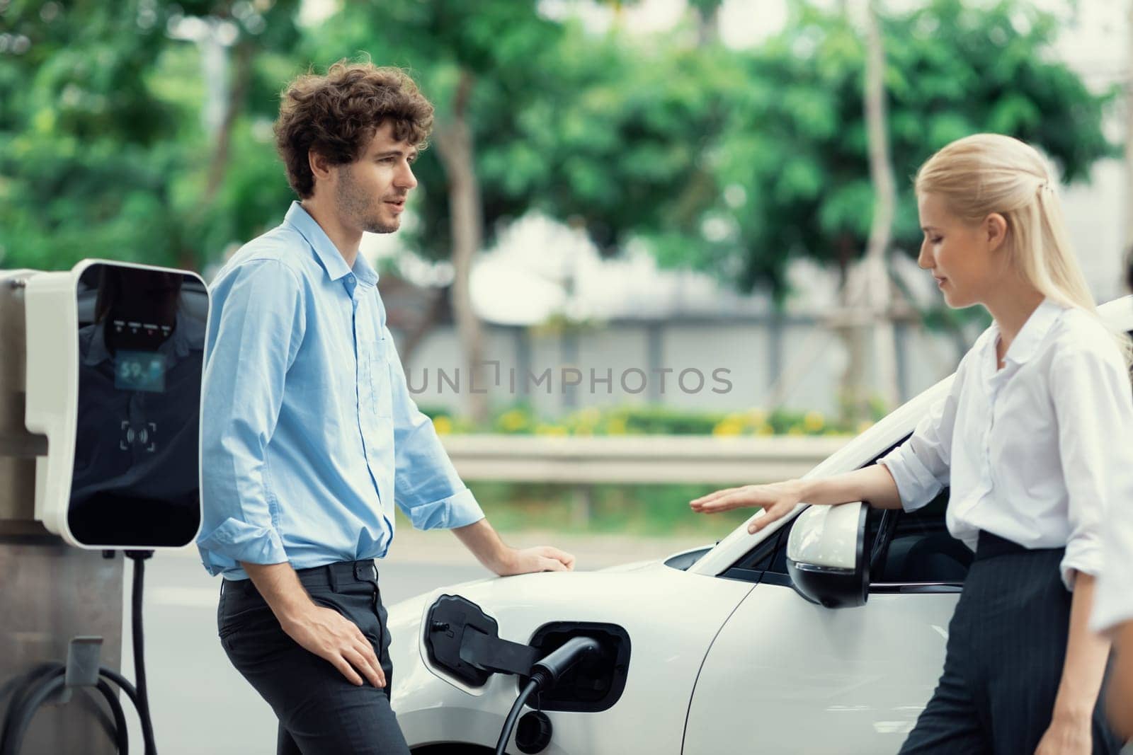 Progressive businessman and businesswoman with electric car parking and connected to public charging station before driving around city center. Eco friendly rechargeable car powered by clean energy.