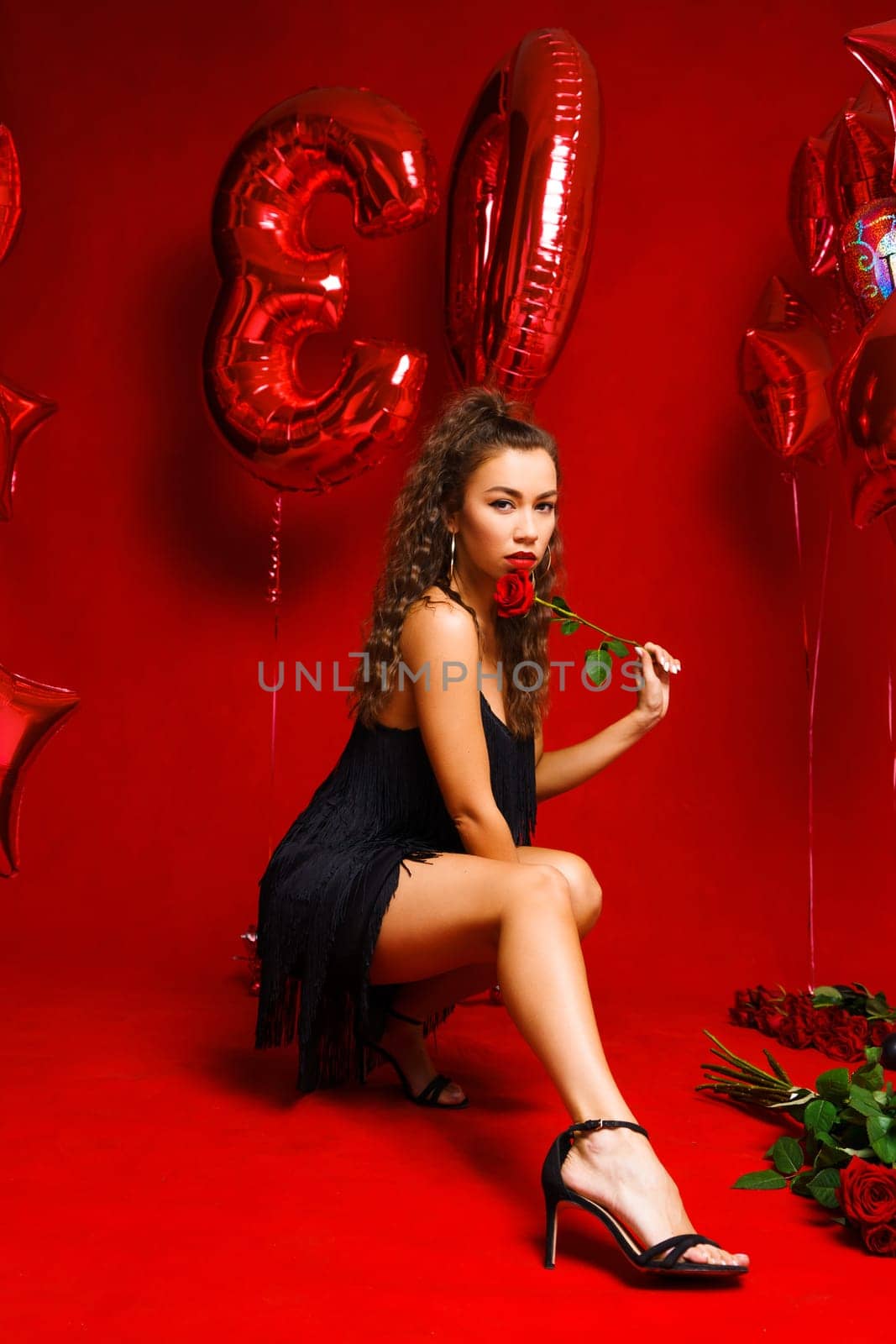 Girl posing sitting on a red background with roses and a bottle of champagne. A young woman of Caucasian ethnicity in a black dress sits on a background with red balloons and cute smiles