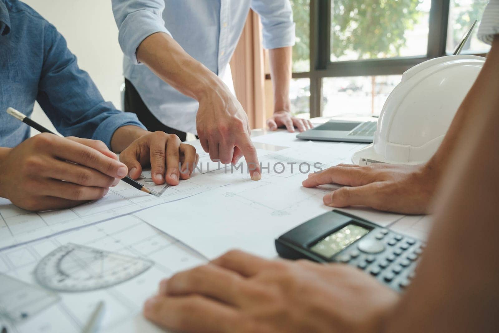 Close-up hands shot of three engineer, developers, or architects working together, drawing graphic design of construction project, and discussing for improvement at desk with papers, compass, helmet, calculator, rulers, and laptop. Engineering and Teamwork concept.