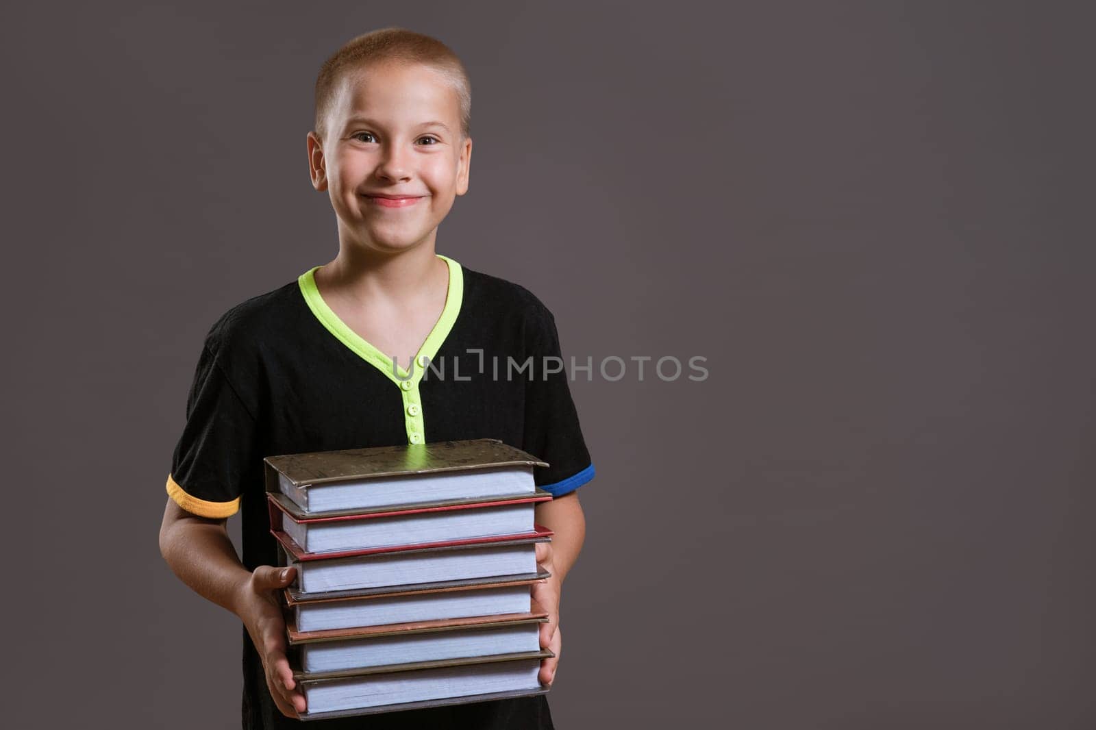 Cheerful caucasian boy in a black t-shirt holds a stack of books on a gray background. Education concept