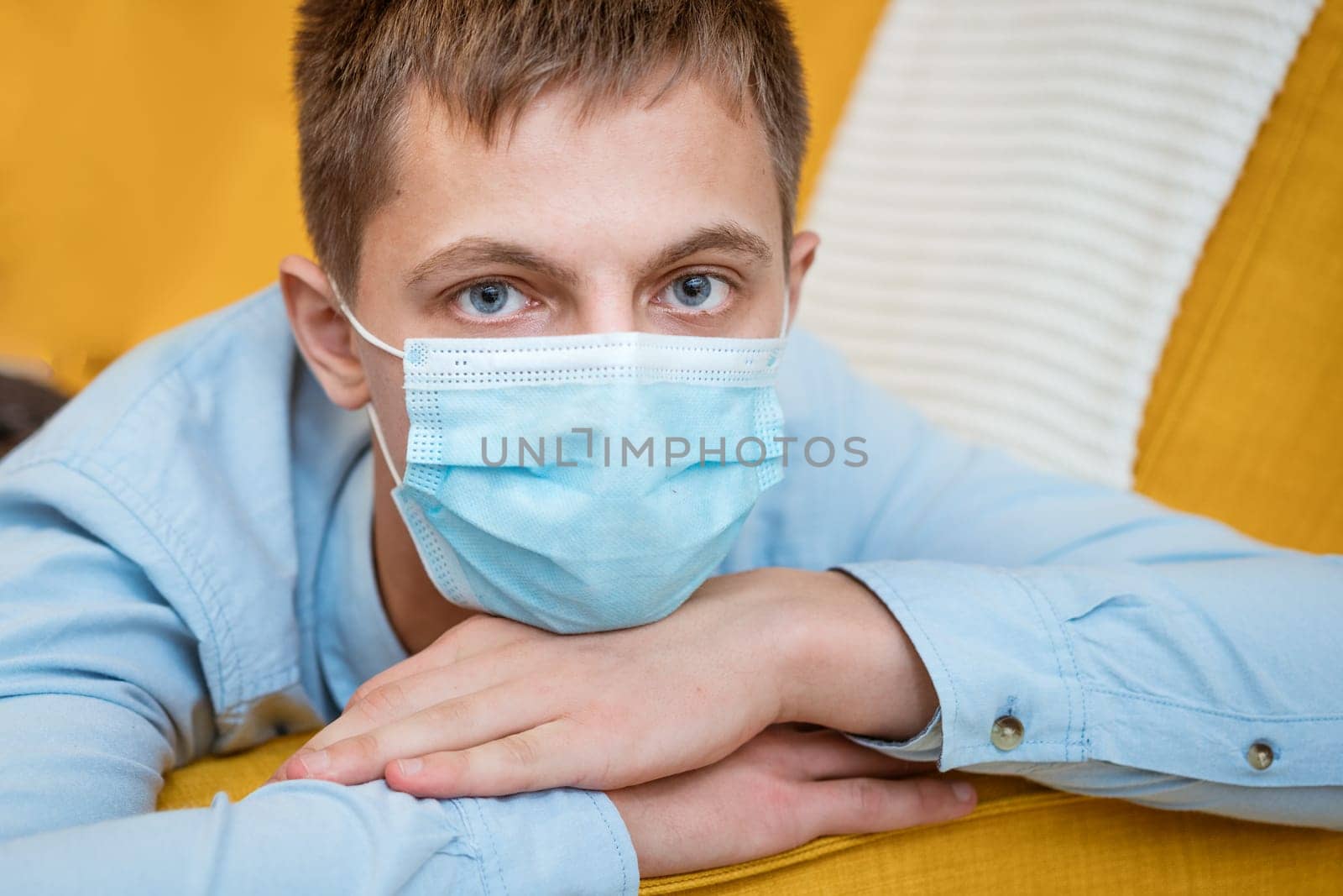 a young guy of Caucasian ethnicity in a shirt and a protective mask sits on the couch alone at home. Concept of being at home during global quarantine