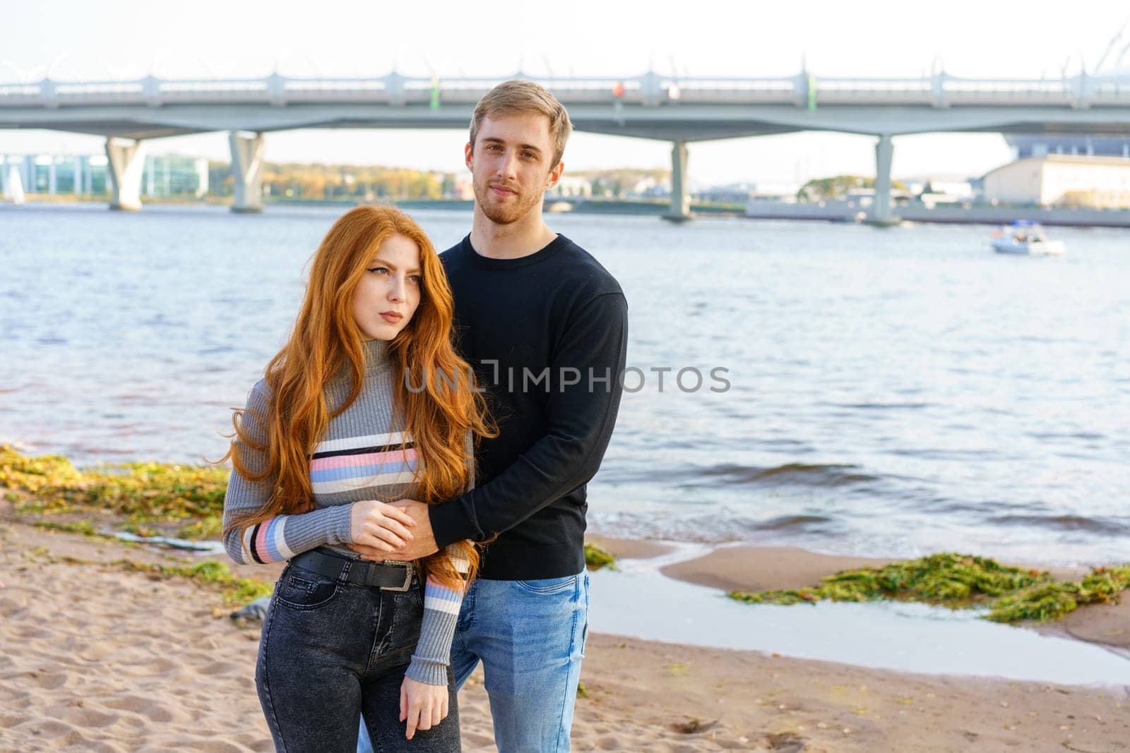 Young couple of man and woman with long red hair of Caucasian ethnicity, in casual clothes, stand on the bank of the river embracing happy on a summer day against the background of the cityscape