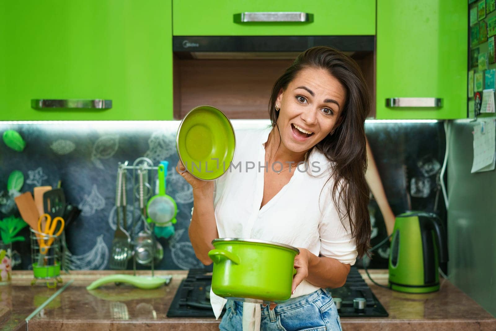 A young woman is holding a saucepan and smiling against the background of a kitchen. Caucasian housewife cheerfully opens the pot lid in casual clothes. Cheerful girl preparing food