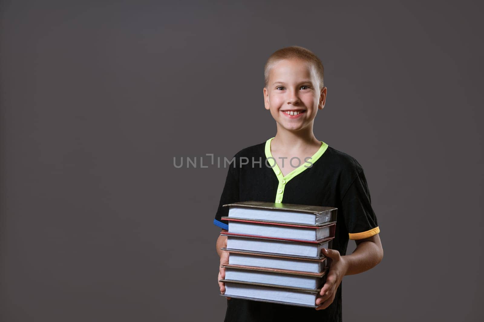 Cheerful caucasian boy in a black t-shirt holds a stack of books on a gray background. Education concept