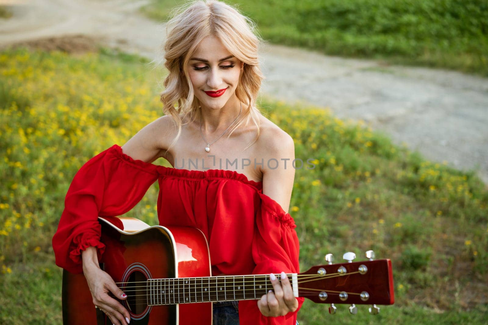 The woman in red is sitting on the green grass with a red guitar. Girl musician plays the guitar outdoors. Blonde caucasian appearance