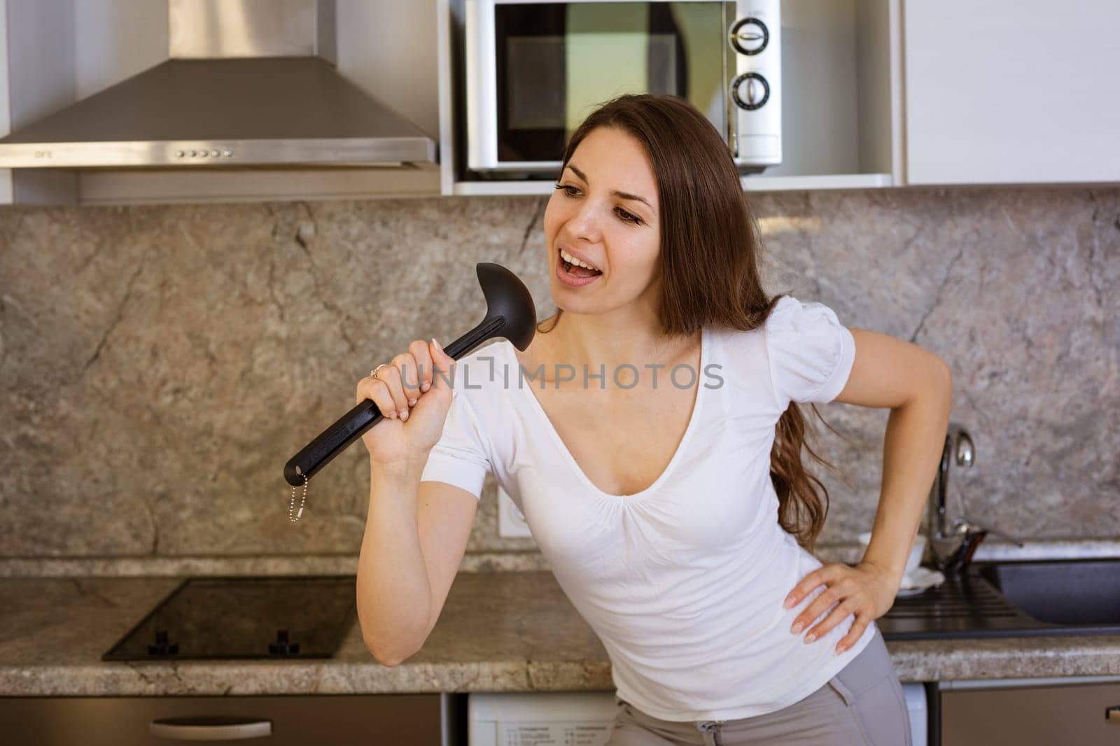 a young brunette woman in a white T-shirt sings in a modern kitchen with a ladle, using it as a microphone