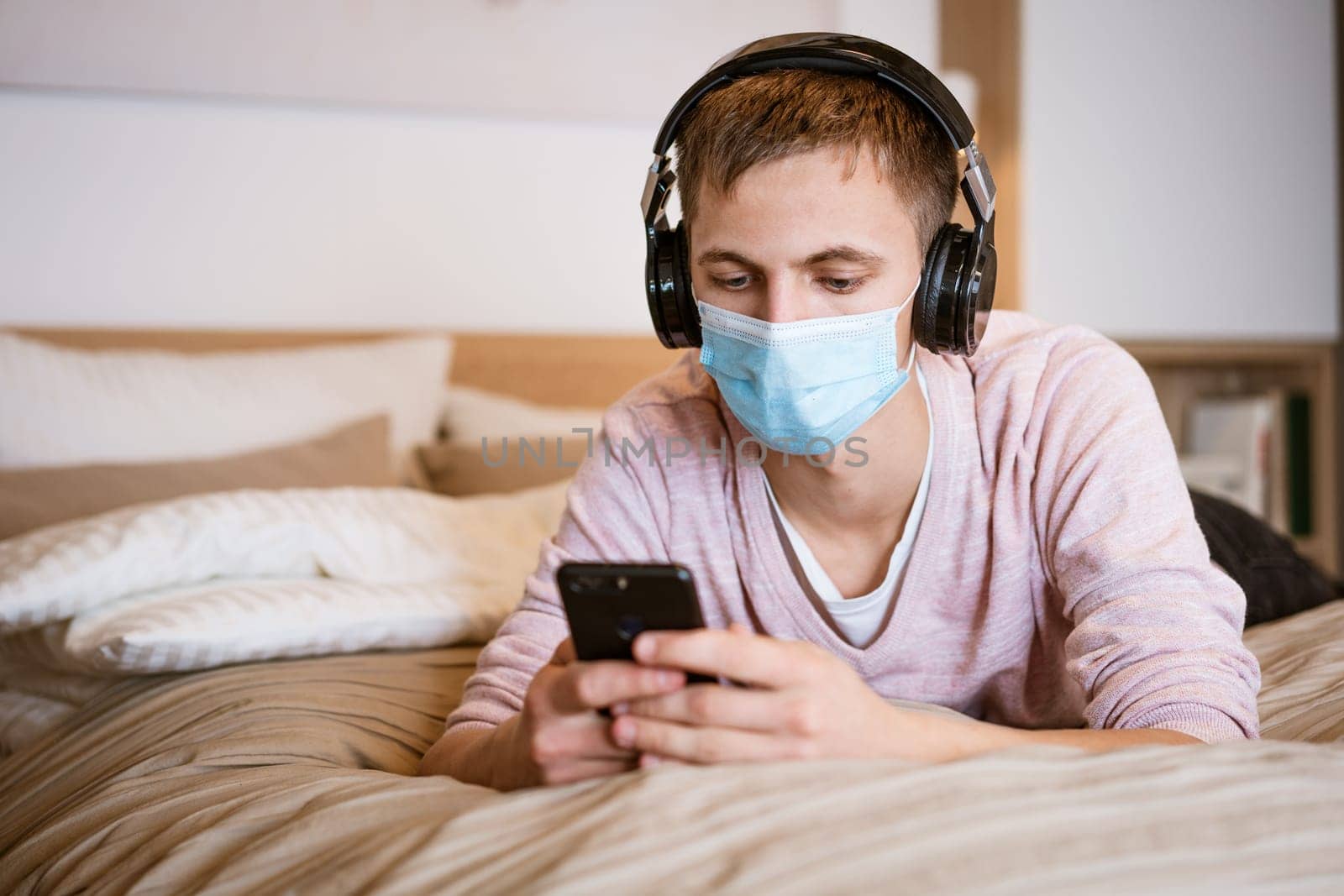 a guy of Caucasian nationality in casual clothes, wearing headphones on the bed in a protective medical mask with a phone in his hand, watching a video online on a Wi-Fi network