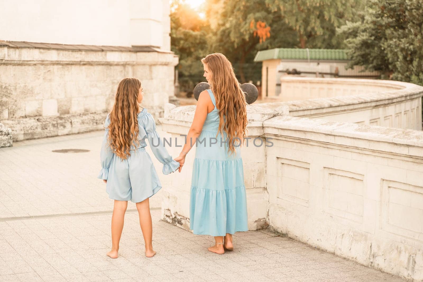 Daughter mother holding hands. In blue dresses with flowing long hair against the backdrop of a sunset and a white building. by Matiunina