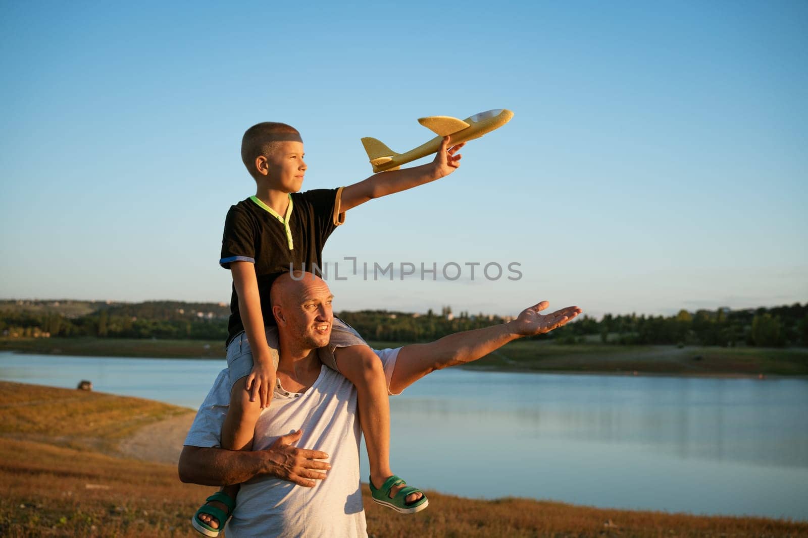 Happy father holding son on shoulders at sunset with airplane in hand, concept of relationship with children