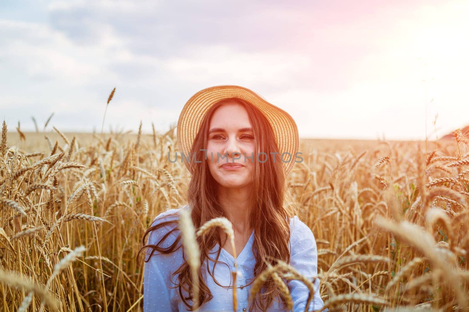 Happy girl sits in wheat field in straw hat. Cute young woman of Caucasian ethnicity in casual clothes enjoys ripe golden wheat in a field. Free woman concept