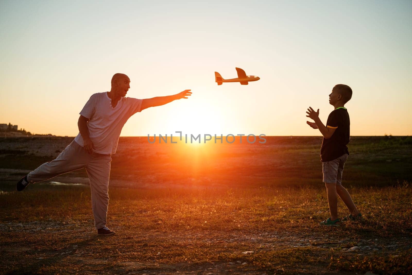 Soft focus of father and son playing toy airplane in meadow at sunset with happy emotions. Family, vacation and travel concept. At sunset in summer they launch an airplane against background sky