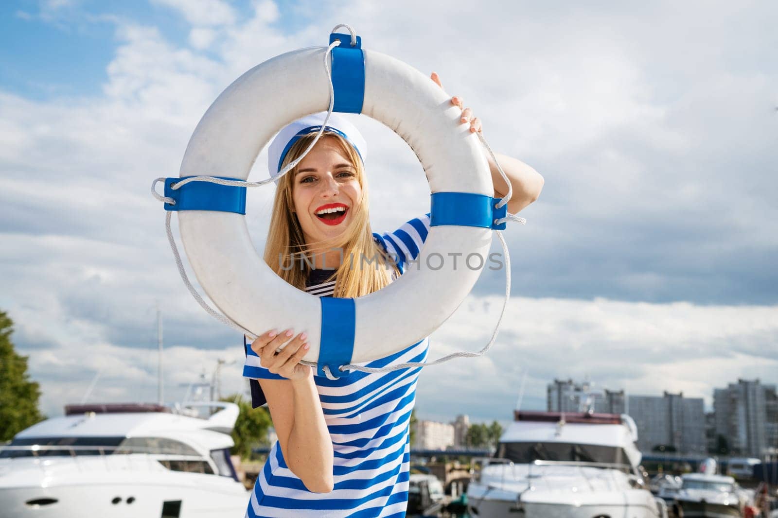 Young happy woman of Caucasian appearance in a blue striped dress standing on a yacht posing with a lifebuoy in her hand, against the background of a blue sky with clouds on a summer sunny day