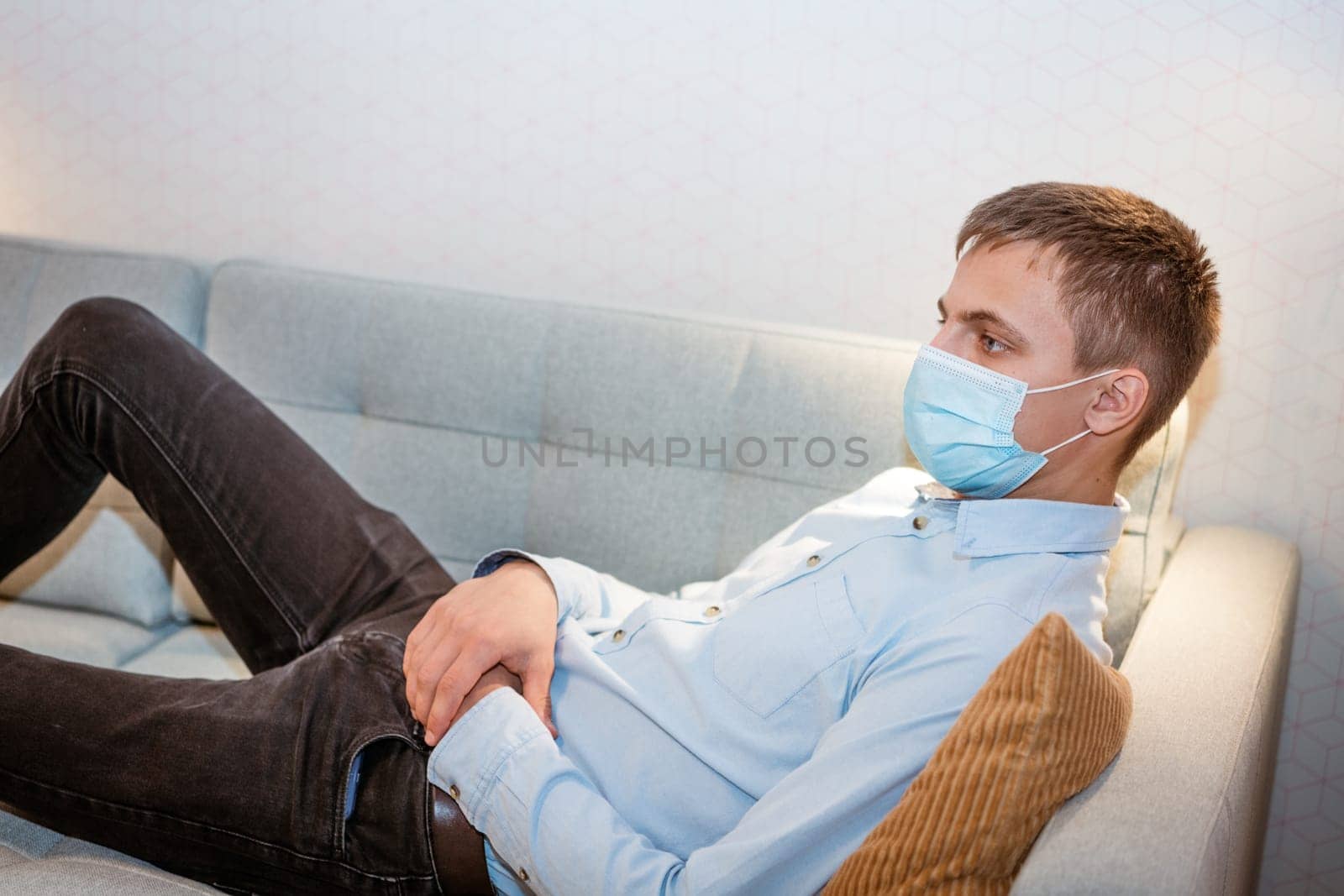 a young guy of Caucasian ethnicity in a shirt and a protective mask sits on the couch alone at home. Concept of being at home during global quarantine