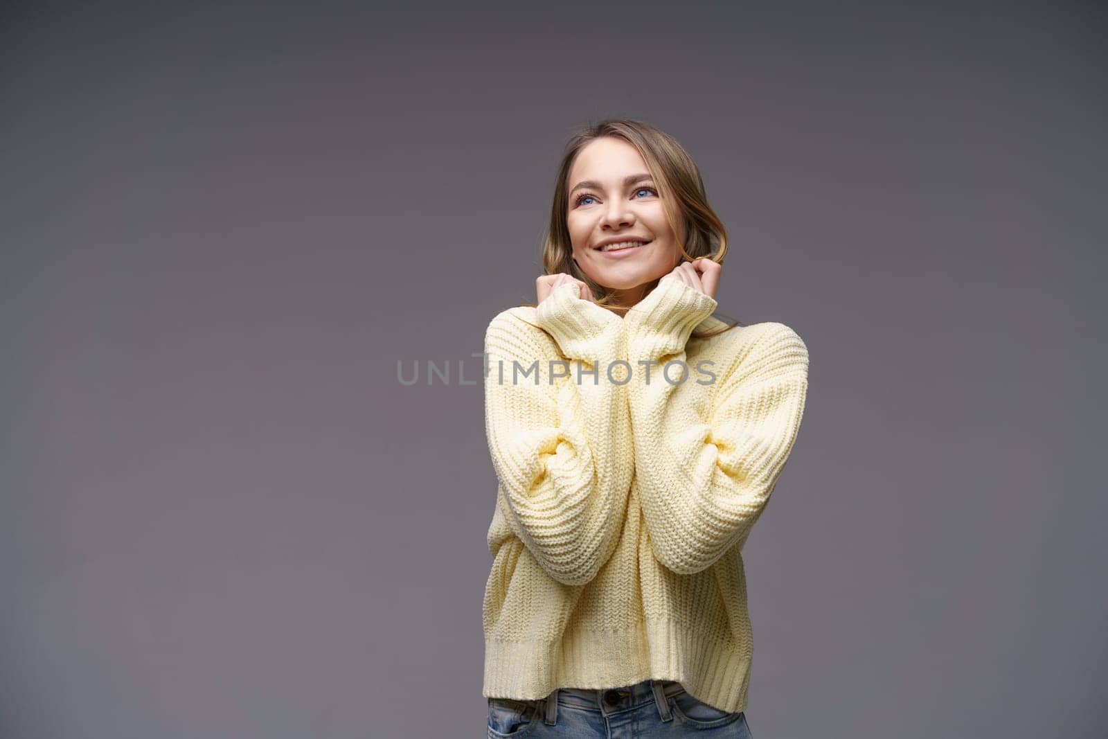 Portrait of a beautiful young girl of Caucasian ethnicity in a yellow sweater on a gray background cute smiling. Keeps his hands to his face.