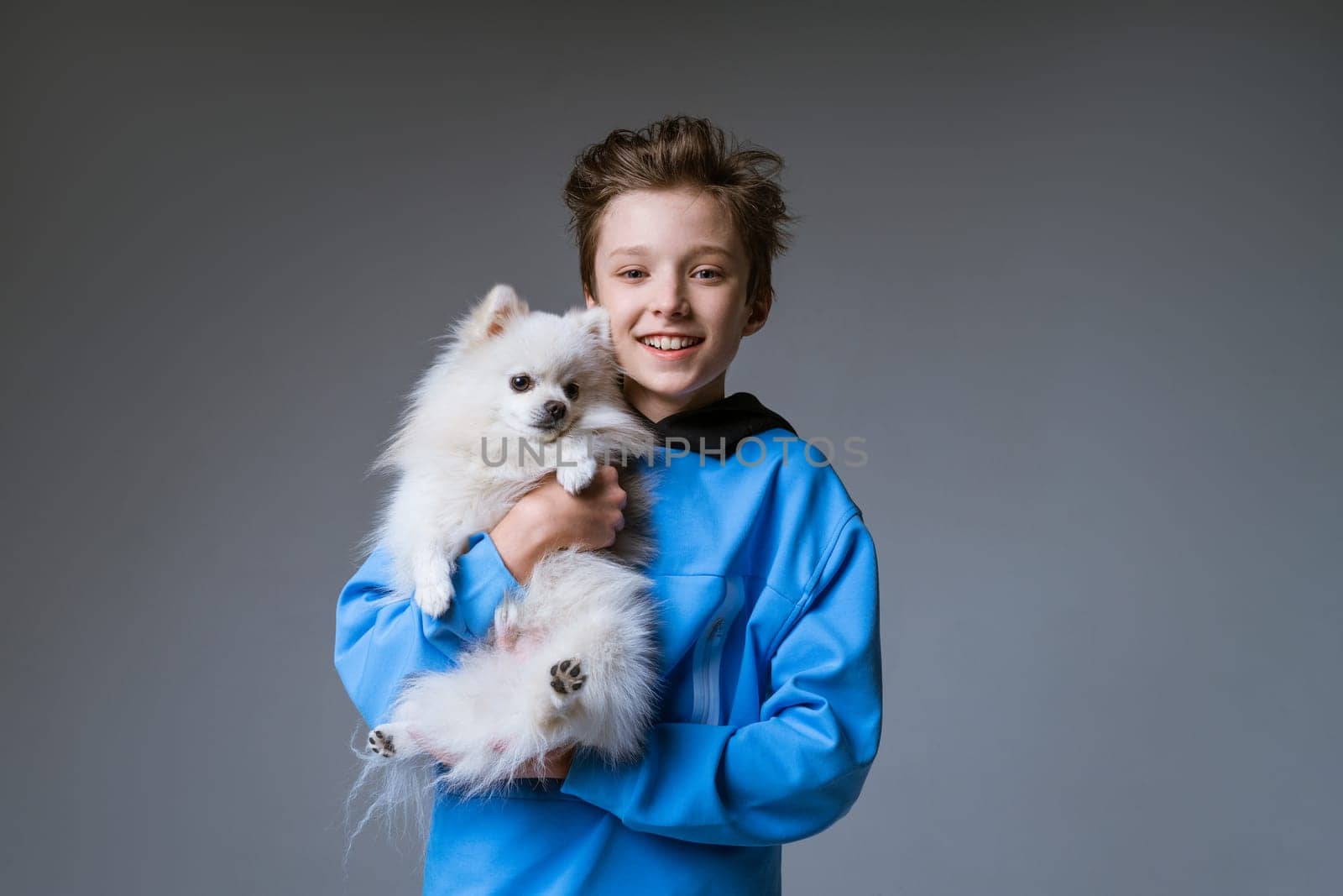 happy boy blond European appearance in blue clothes, holding a white dog on a gray background and smiling with snow-white teeth