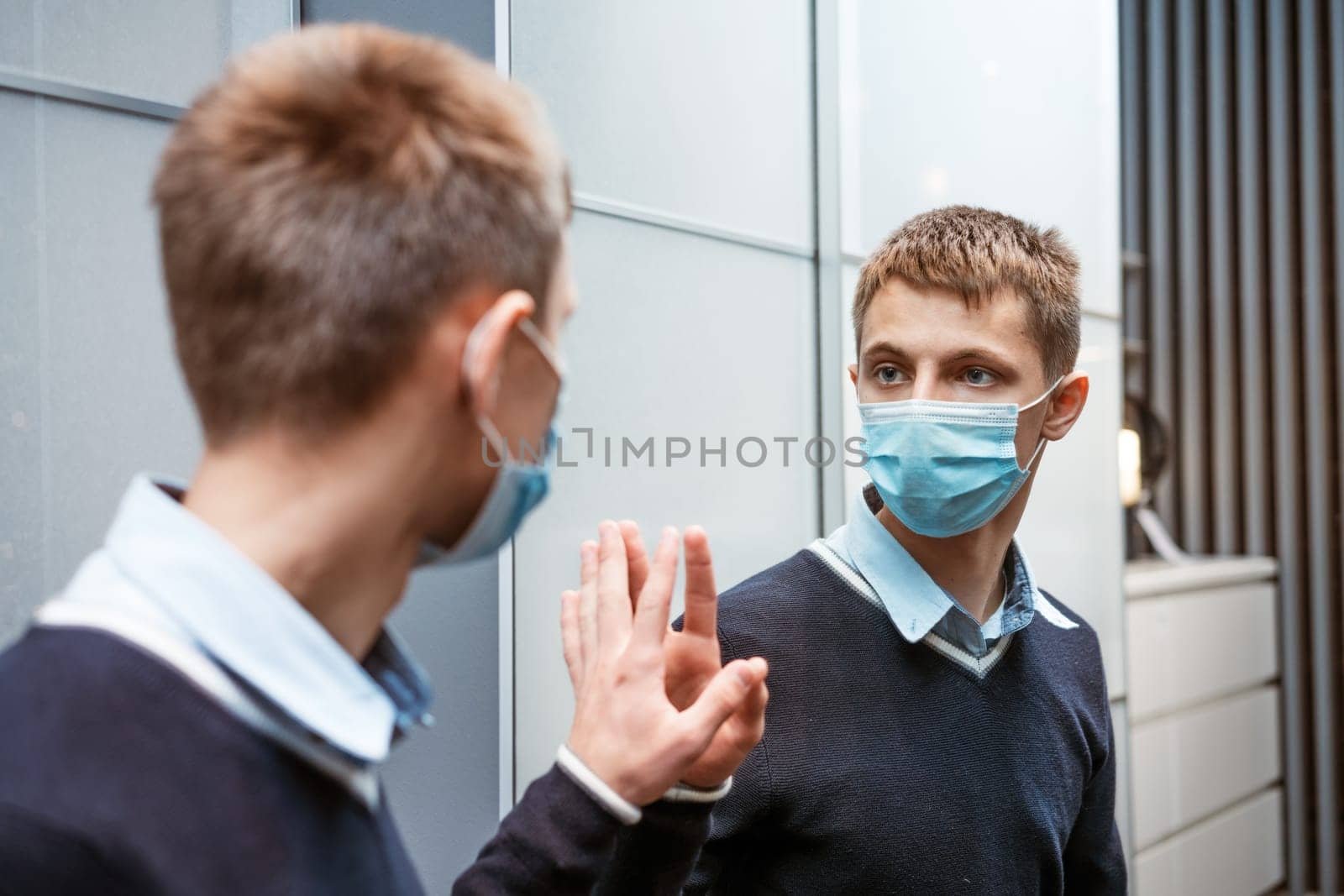 young caucasian man in a protective mask looks in the mirror, adjusts the medical mask against the virus. The concept of wave ipidemii