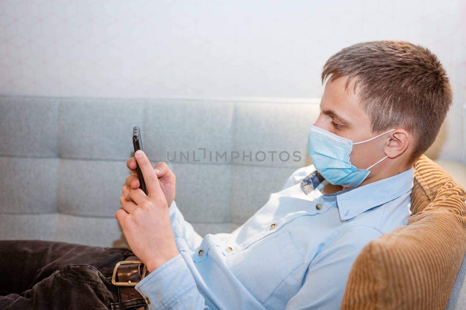 a young man sits on a sofa wearing a protective mask with a phone in his hand by EkaterinaPereslavtseva