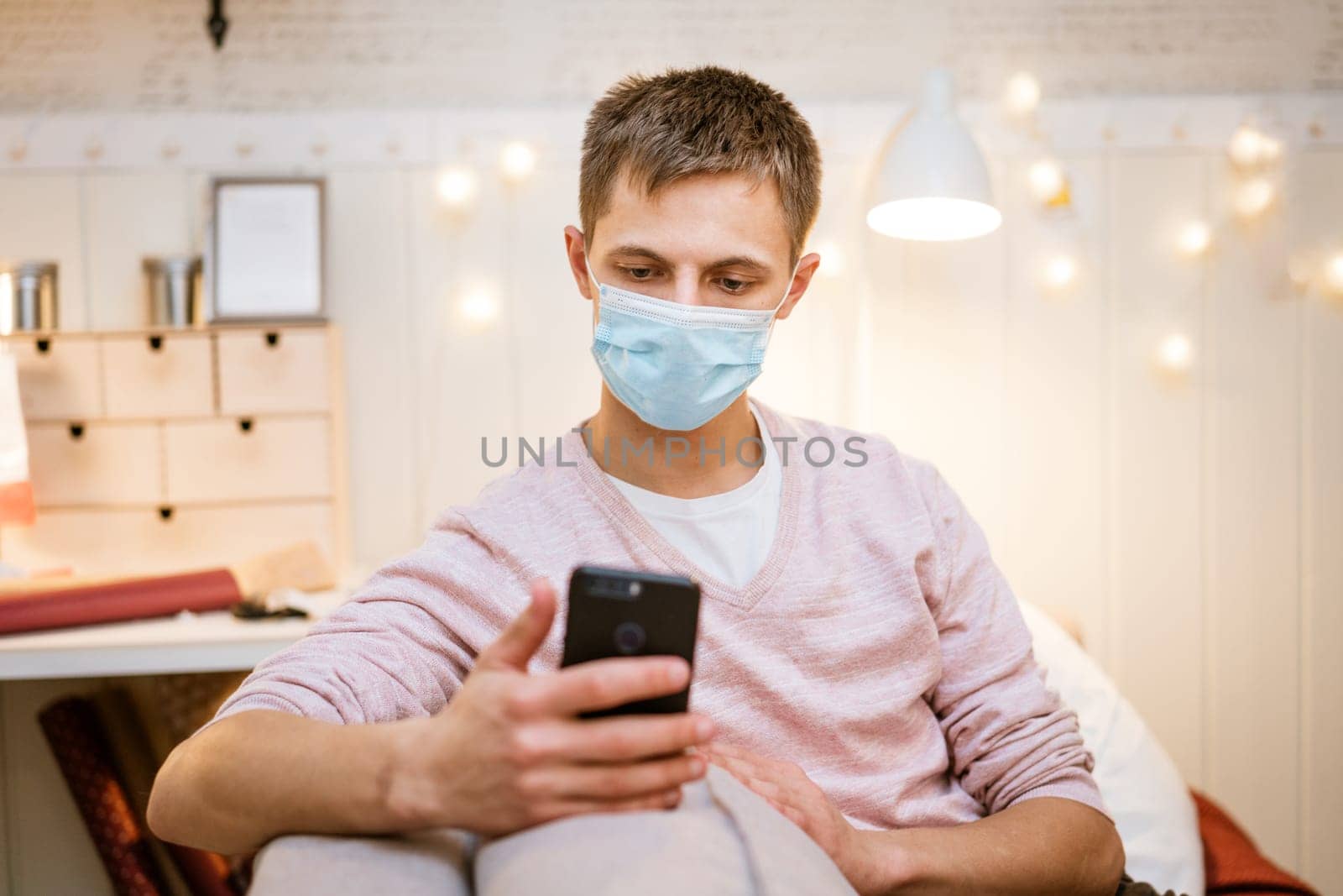 young man of European appearance in casual clothes, at home on the couch in a medical mask communicates by video communication on the phone, the concept of remote online communication on the Internet