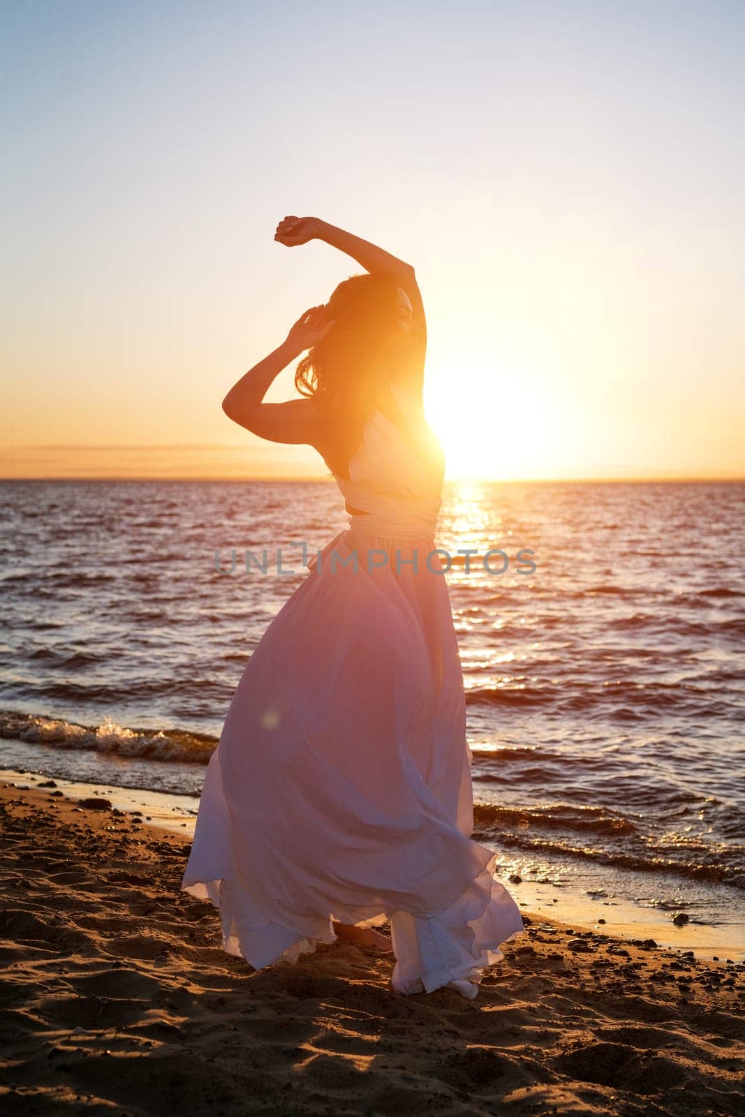A beautiful young woman of Caucasian appearance with dyed hair posing on the seashore in the sun at sunset in a white dress in the wind. Romantic slender girl in nature enjoys relaxation