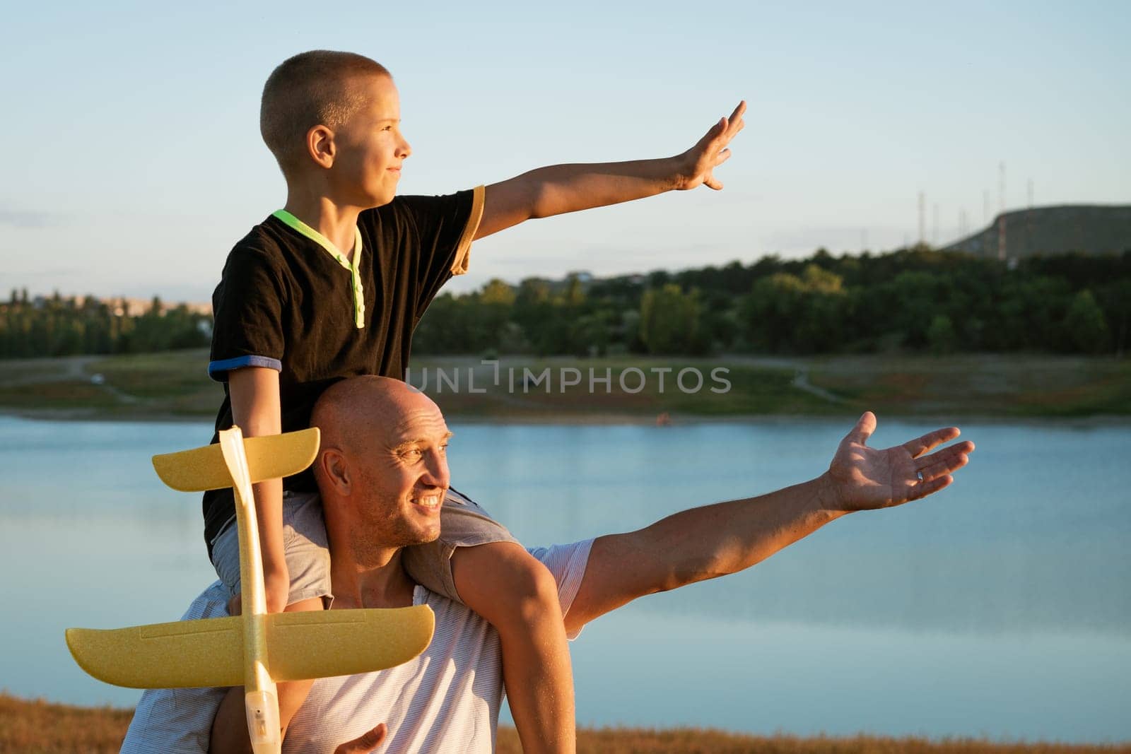 Happy father holding son on shoulders at sunset with airplane in hand, concept of relationship with children