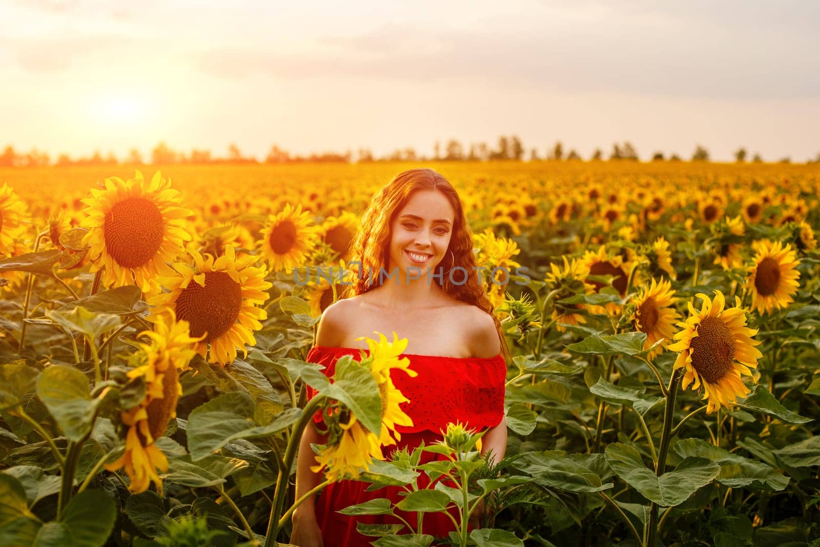 Young woman smiling at sunset in a field of sunflowers. A cute girl of Caucasian appearance in a red dress is happy and free to stand in the evening in the rays. Natural beauty in nature