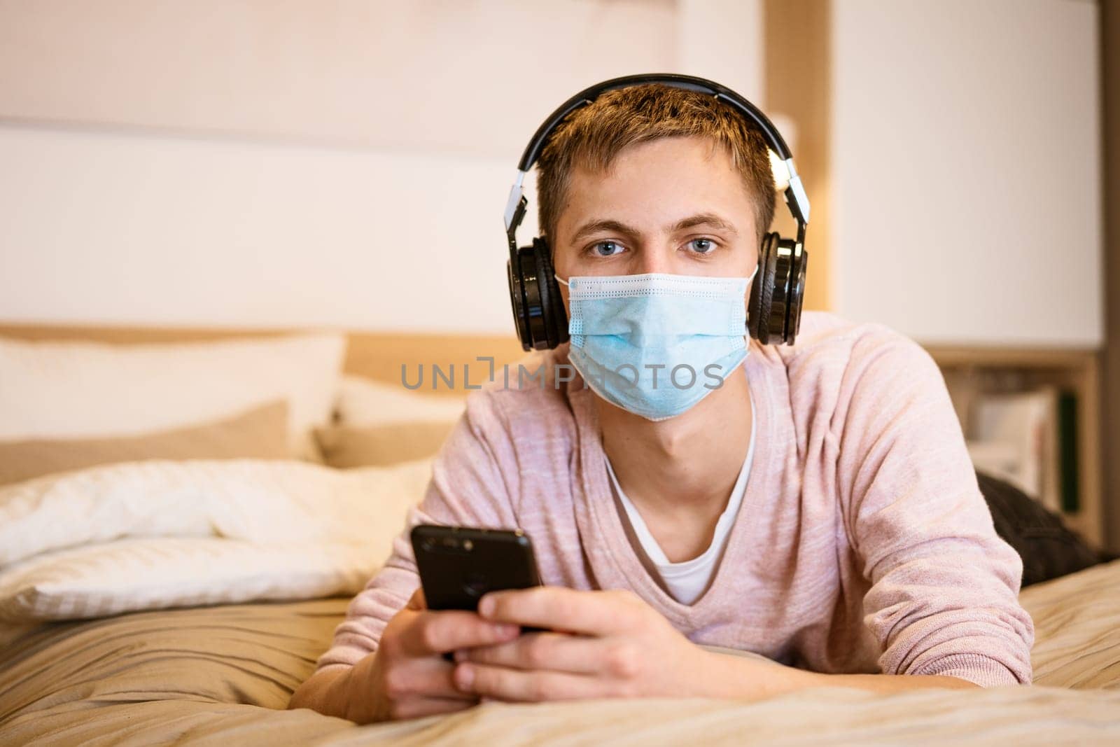 man of European appearance in casual clothes, wearing headphones and protective mask with phone his hands on bed at home, spending time Internet while sitting in quarantine during Lokdan