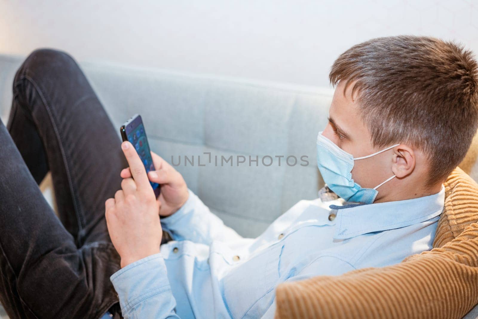 young man of European appearance in casual clothes, at home on the couch in a medical mask communicates by video communication on the phone, the concept of remote online communication on the Internet