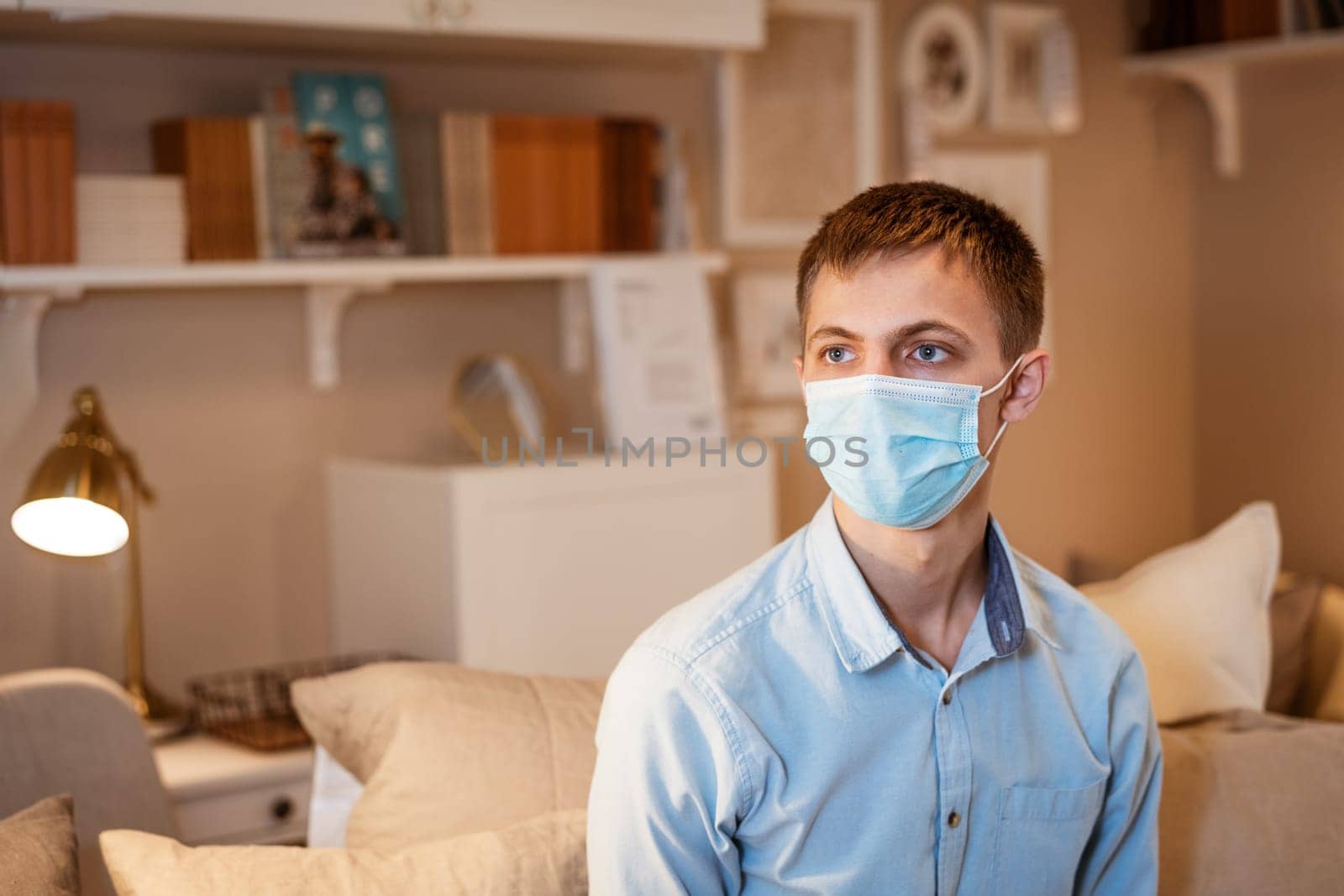 a young guy of Caucasian ethnicity in a shirt and a protective mask sits on the couch alone at home. Concept of being at home during global quarantine