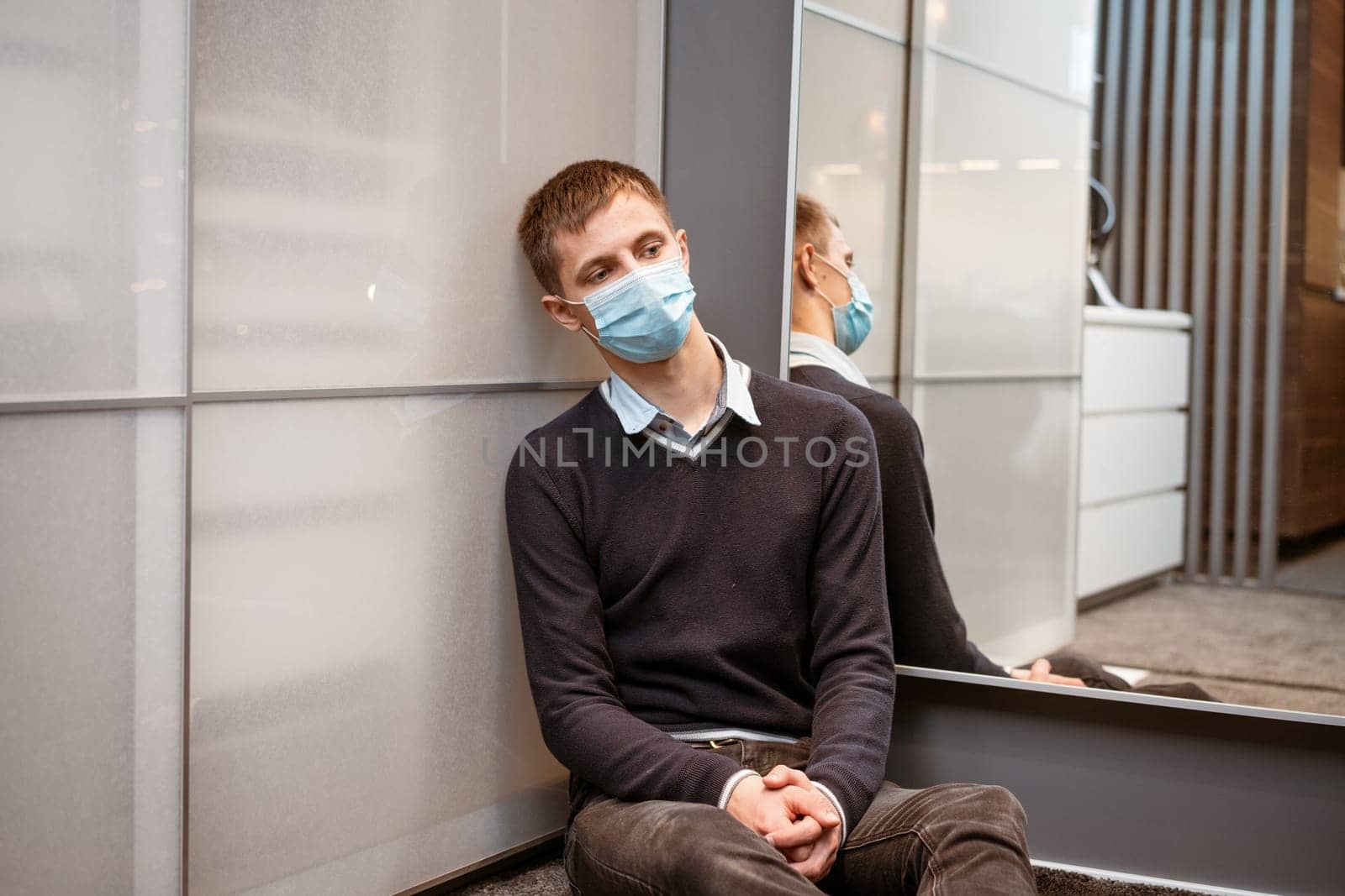 a guy of Caucasian nationality sits in a corner wearing a protective mask sad at the mirror.