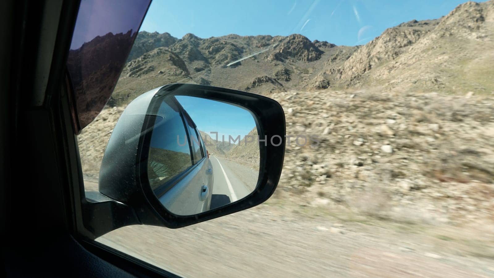 View of the road in the side mirror of the car. Behind there are yellow fences, huge rocky mountains with dry bushes. There are stones lying. Black dusty mirror of a white car. An SUV. Journey.